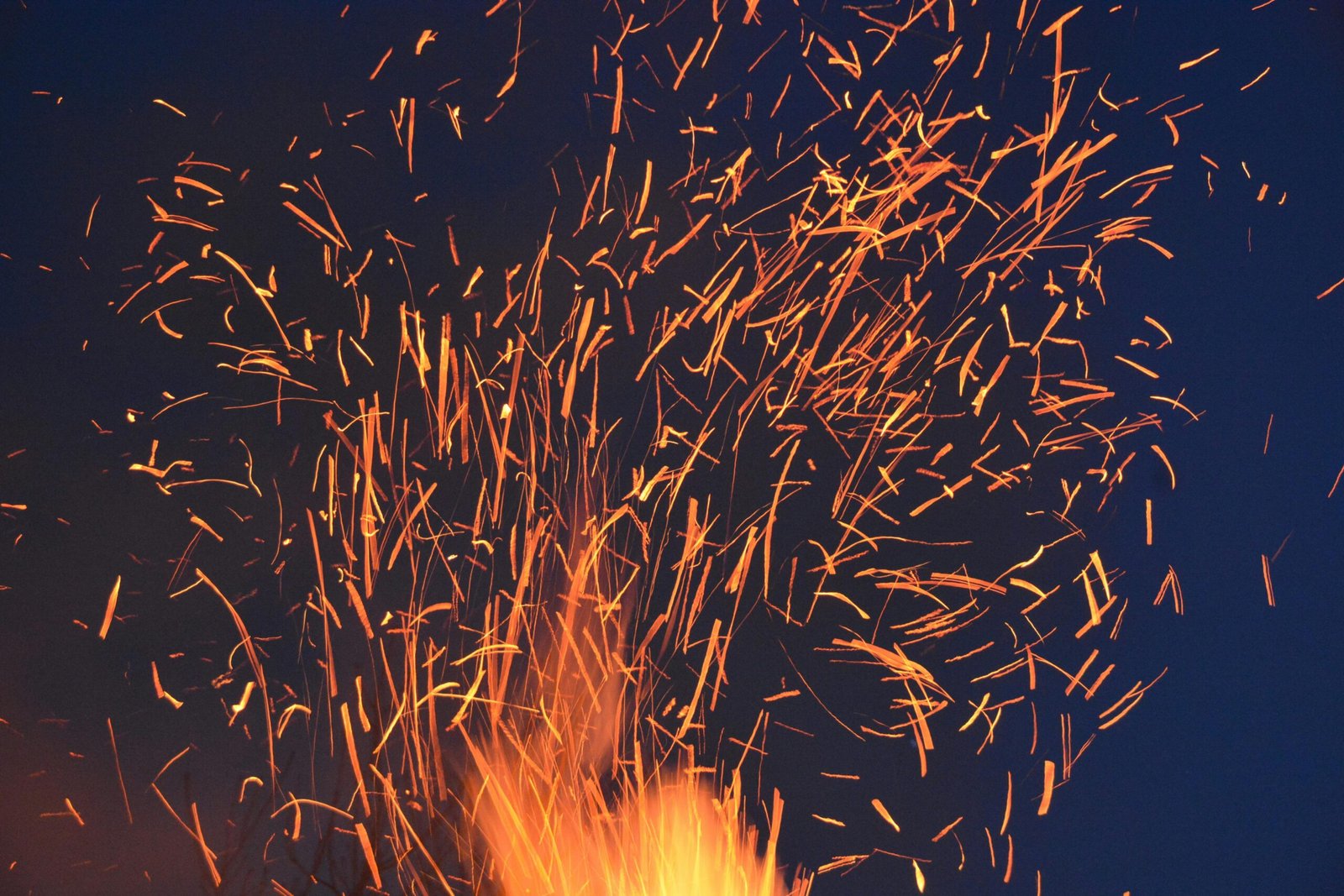 Vibrant orange sparks fly from a fire against a dark blue night sky, creating a dramatic visual.