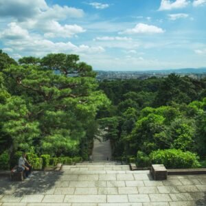 From above of long stairway leading through lush exotic green trees under cloudy blue sky in Fushimi Momoyama Castle park